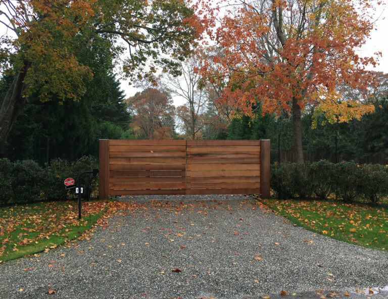A charming wooden gate crafted by East Hampton Fence & Gate stands amid vibrant autumn trees, with fallen leaves carpeting the gravel driveway.